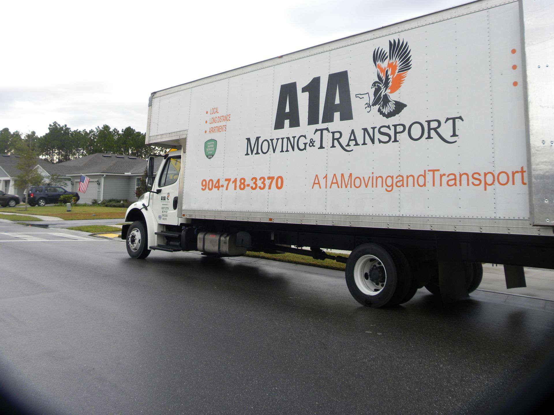 White A1A Moving & Transport truck on a wet road in front of houses; cloudy sky.