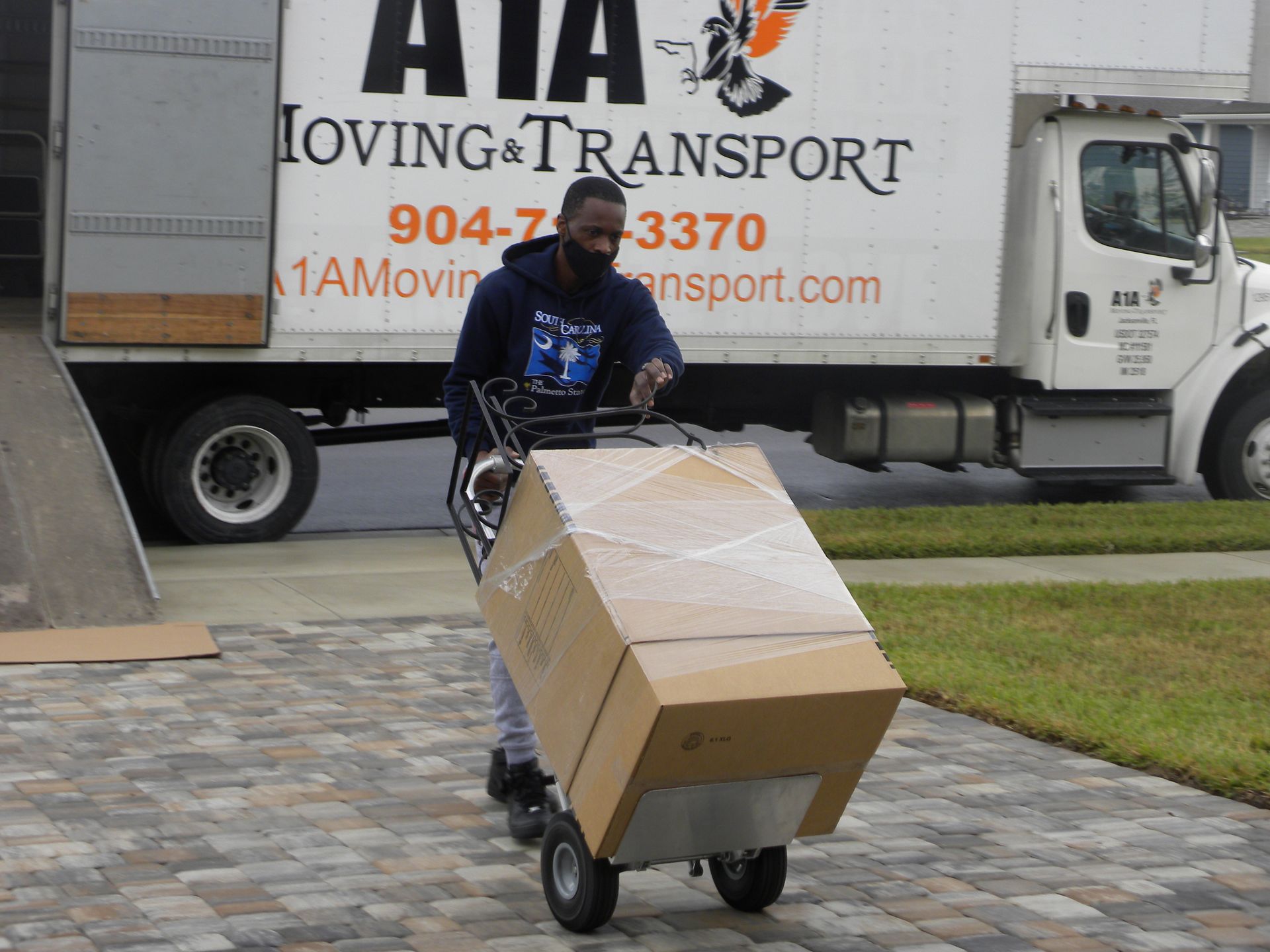 Man in mask moving large box on a dolly to moving truck; parked on a driveway.