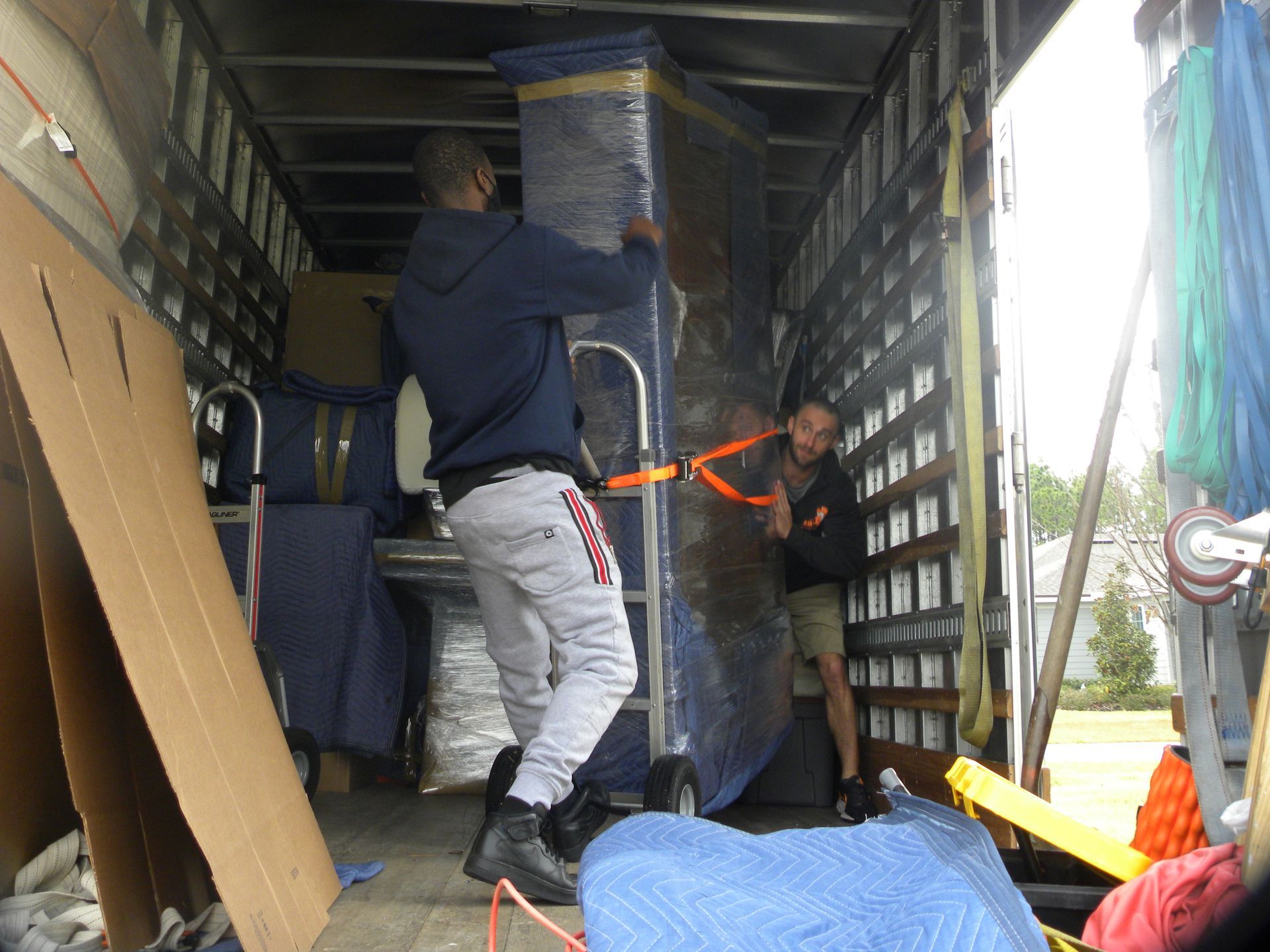 Two people loading a wrapped tall item into a moving truck. The truck's interior is visible.