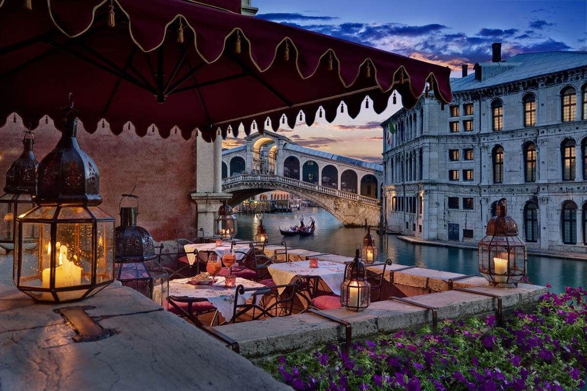 A restaurant with tables and chairs overlooking a canal with a bridge in the background.