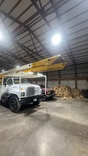 White utility truck with extended yellow boom inside a large warehouse, beside stacked wood chips.