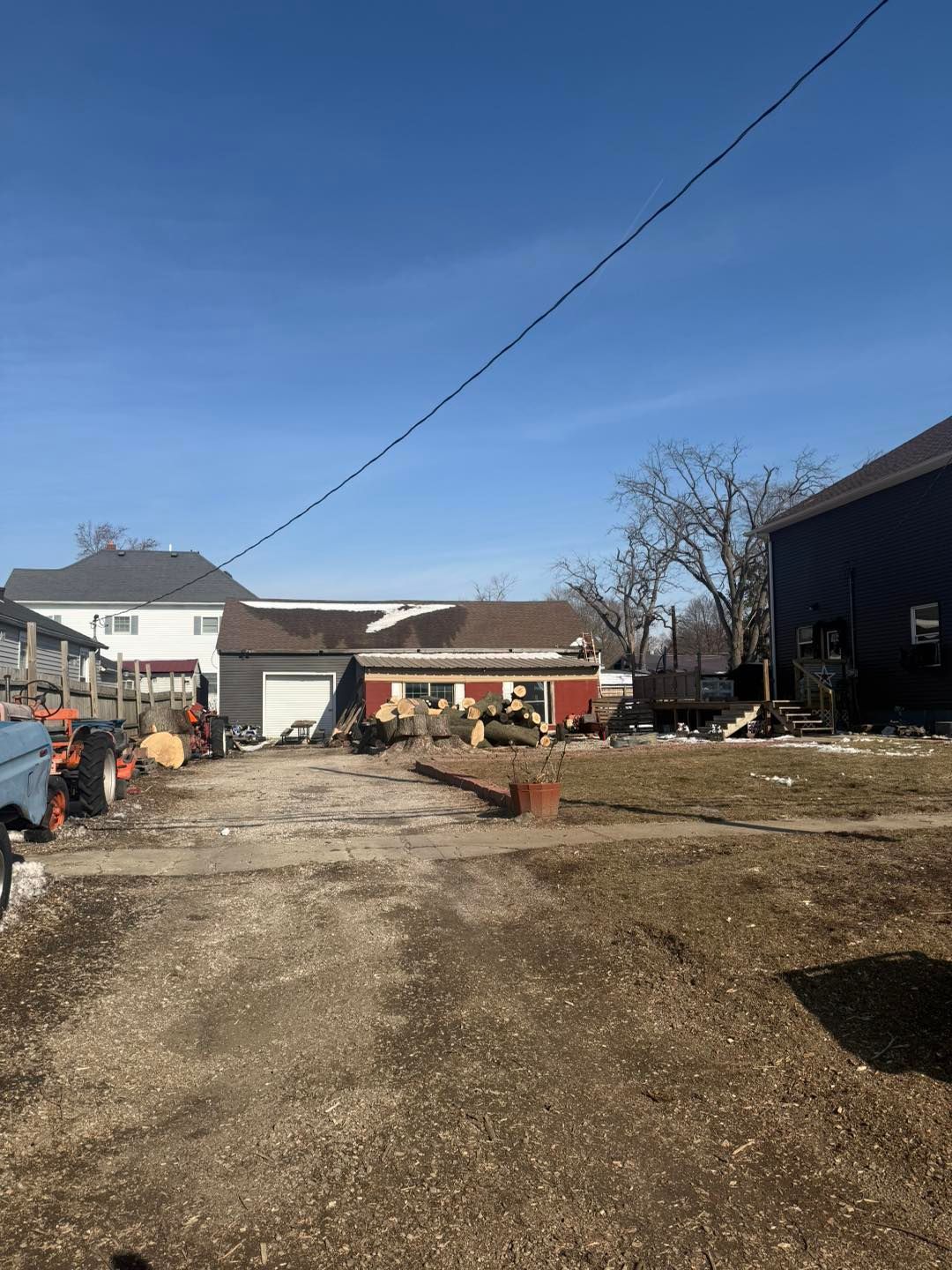 Gravel driveway beside a house and barn on a sunny day, with potted plants and parked equipment.
