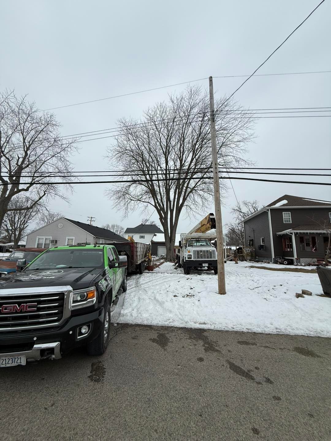 Snow-covered suburban street with parked trucks, bare trees, and houses on an overcast day