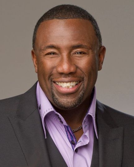 Smiling man in a suit jacket over a purple shirt. Gray background.