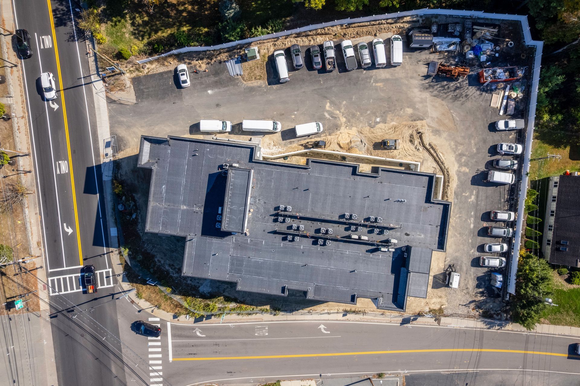 Overhead view of a building with a flat roof, surrounded by cars, construction materials, and a road.