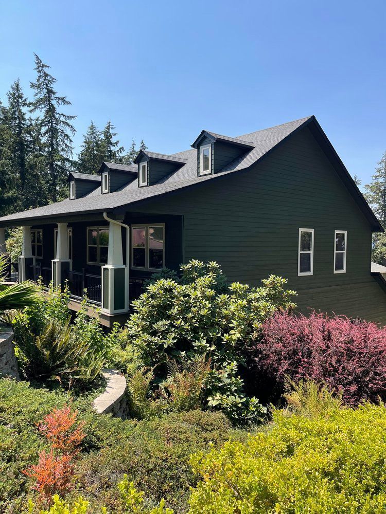 A large green house with a porch is surrounded by bushes and trees.