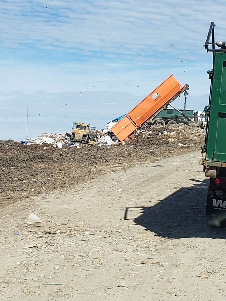 A green garbage truck is driving down a dirt road