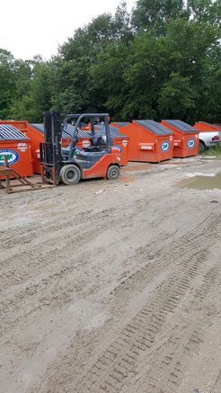 A forklift is driving down a dirt road next to a row of dumpsters.