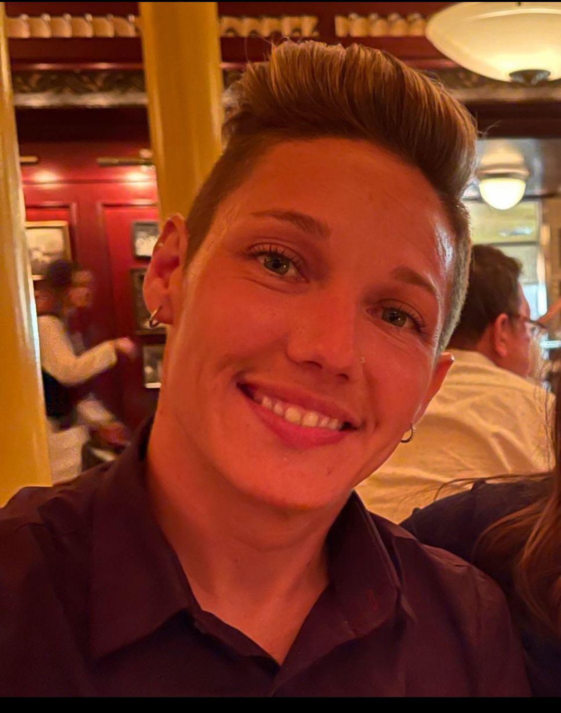 Woman smiling in a restaurant; short, styled hair; wearing a dark purple shirt.