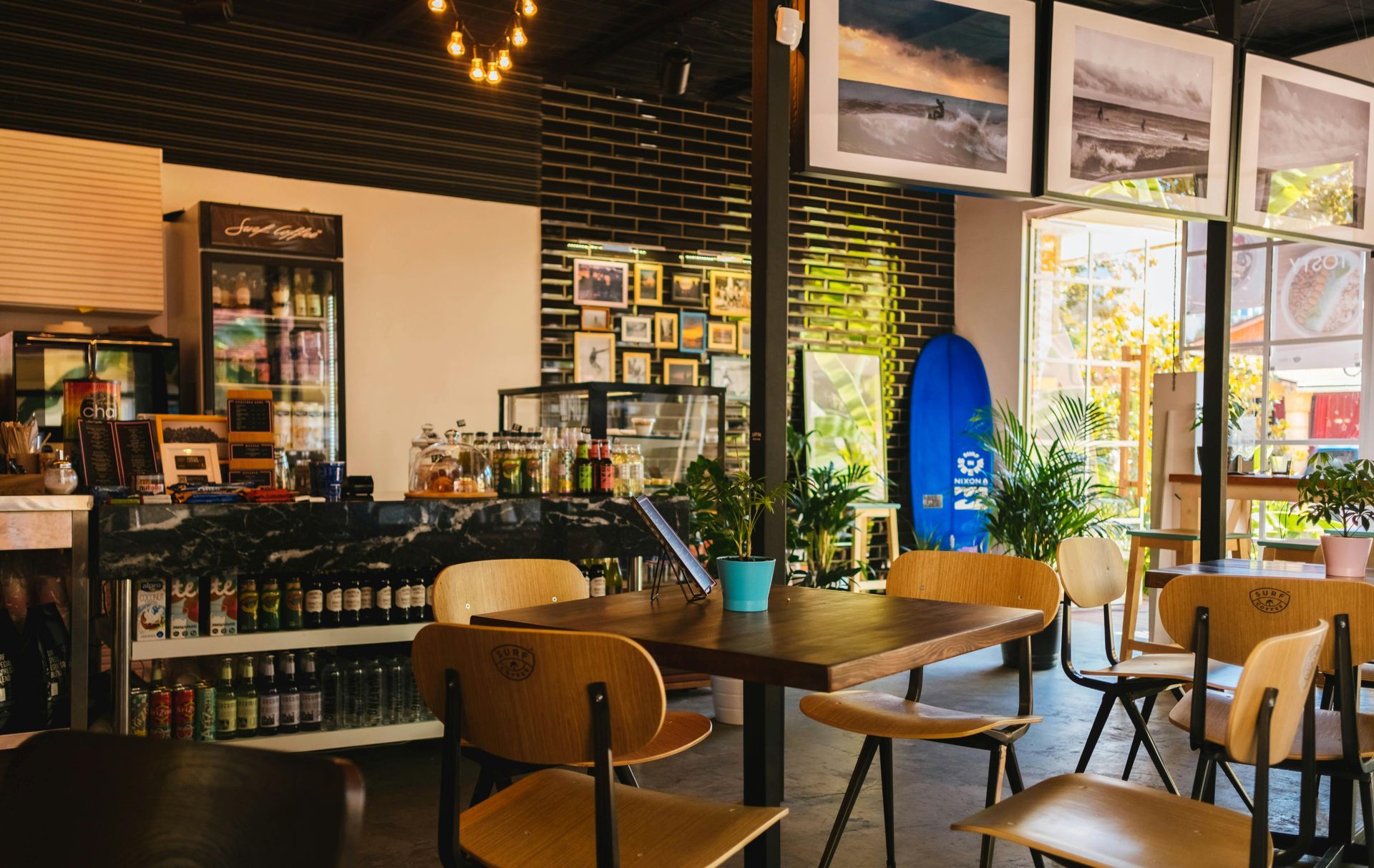 Interior of a cafe with tables and chairs. Surfboard and framed photos on the wall, fridge on the counter.