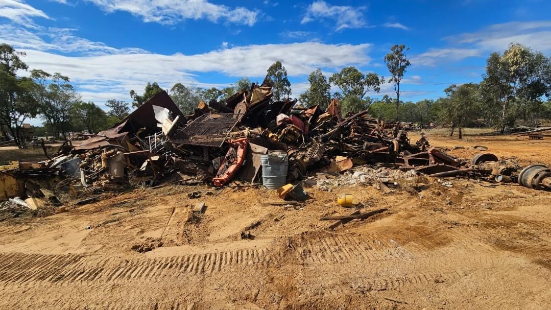A Pile of Charred Metal and Debris on a Sandy Field Under a Blue Sky With Trees — NPX Solutions In Rockhampton City, QLD