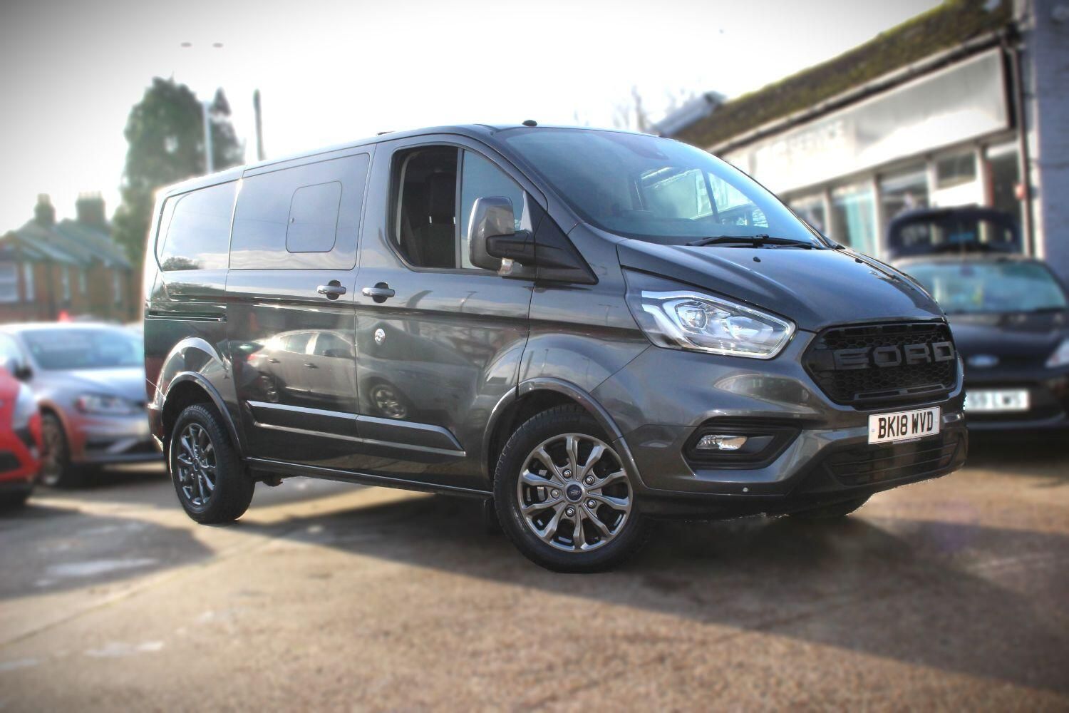 A ford transit custom is parked in front of a car dealership.