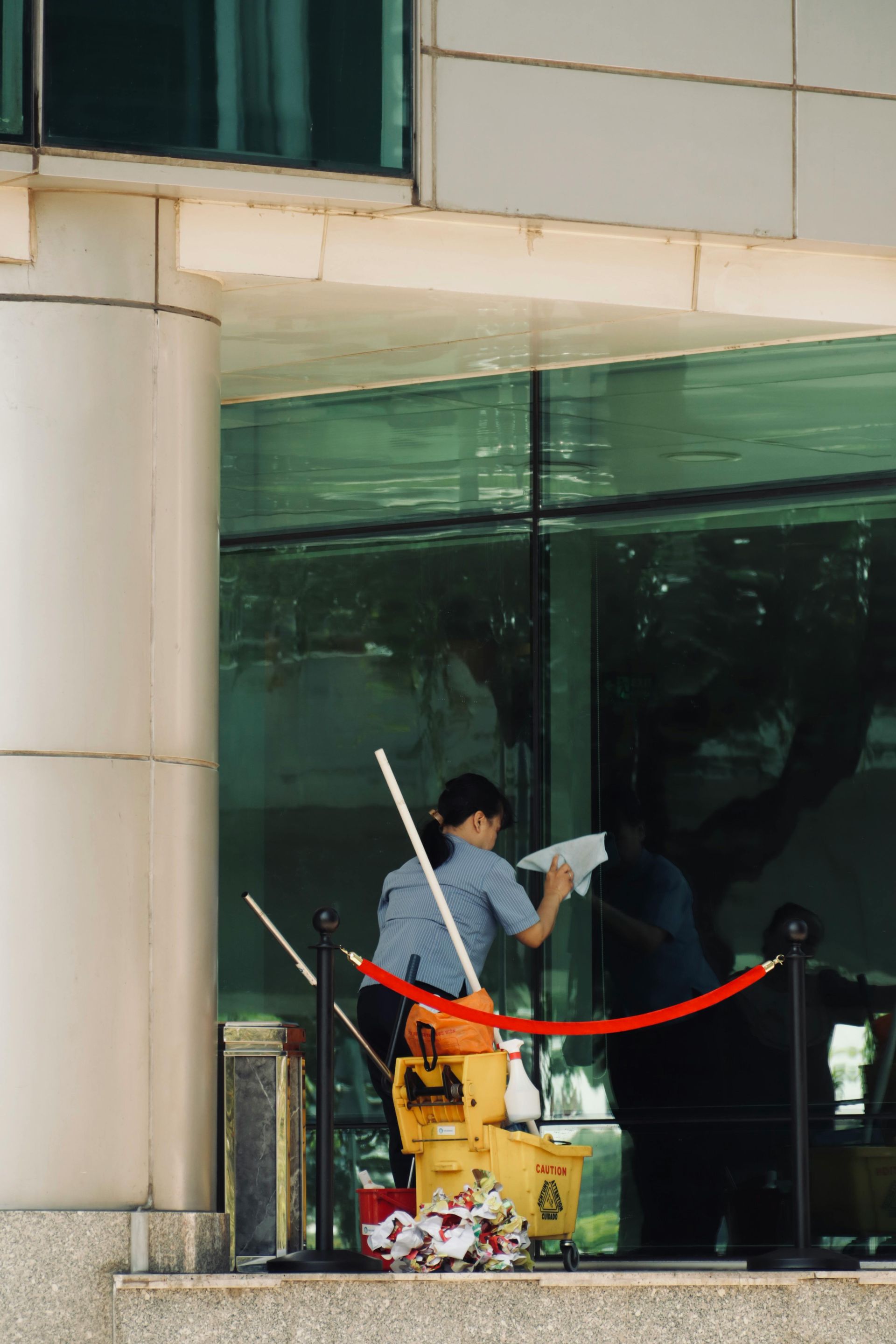 Person in blue uniform cleaning a large green window, standing outside a building with a red rope barrier.