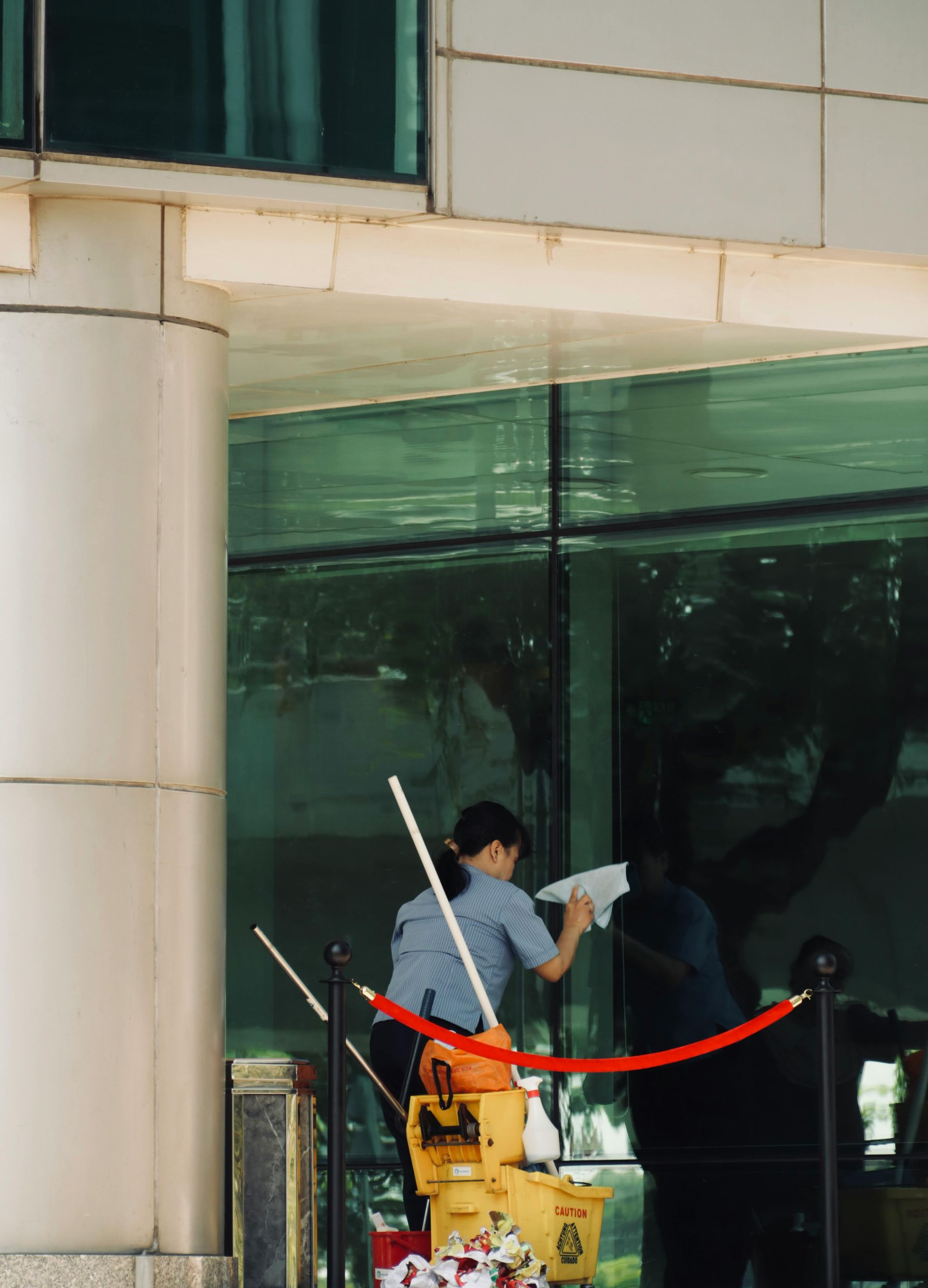 Two people cleaning a glass building exterior; a red rope barrier and cleaning supplies are visible.