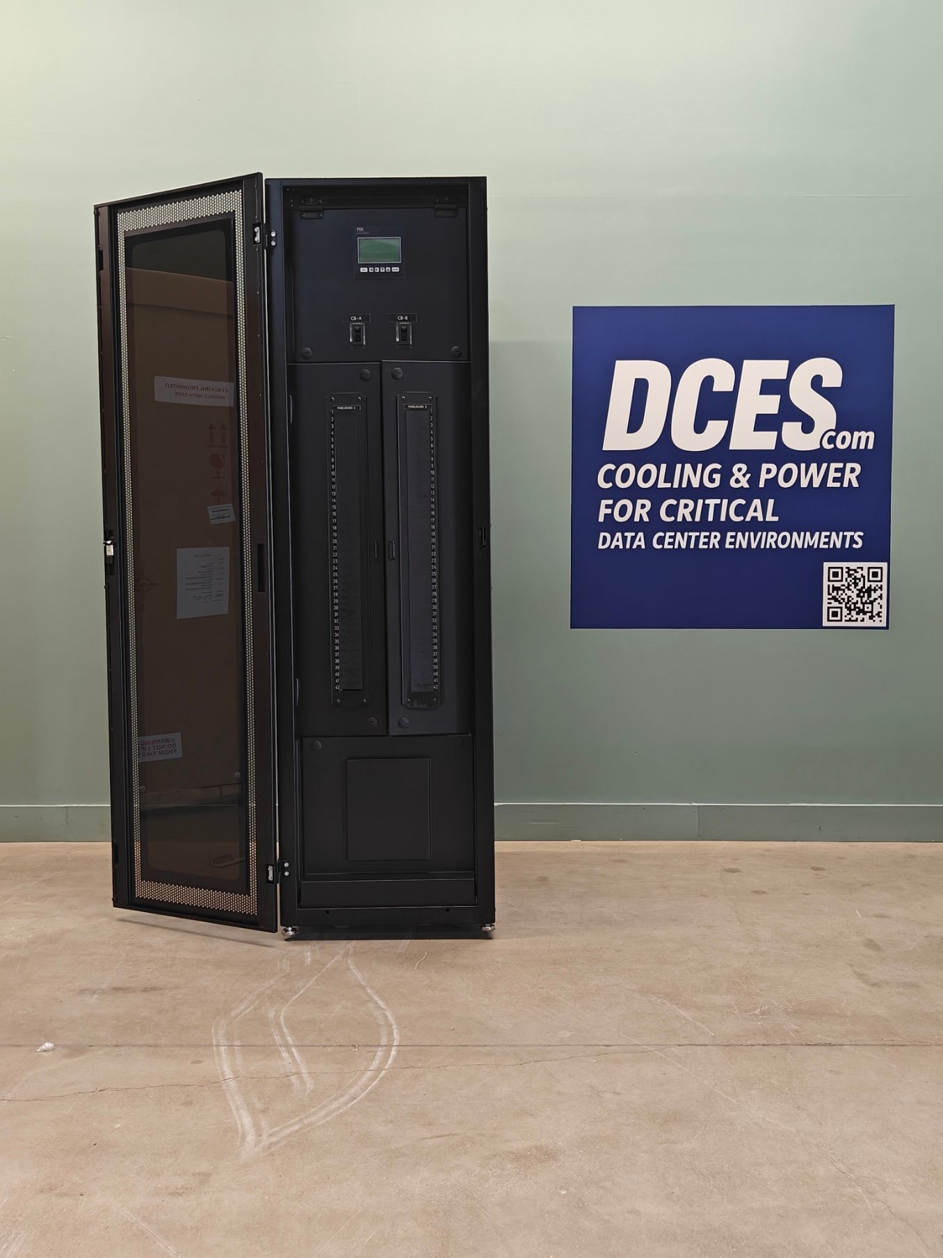 Black server rack with open side door, next to a blue and white DCES sign on a light-colored wall.