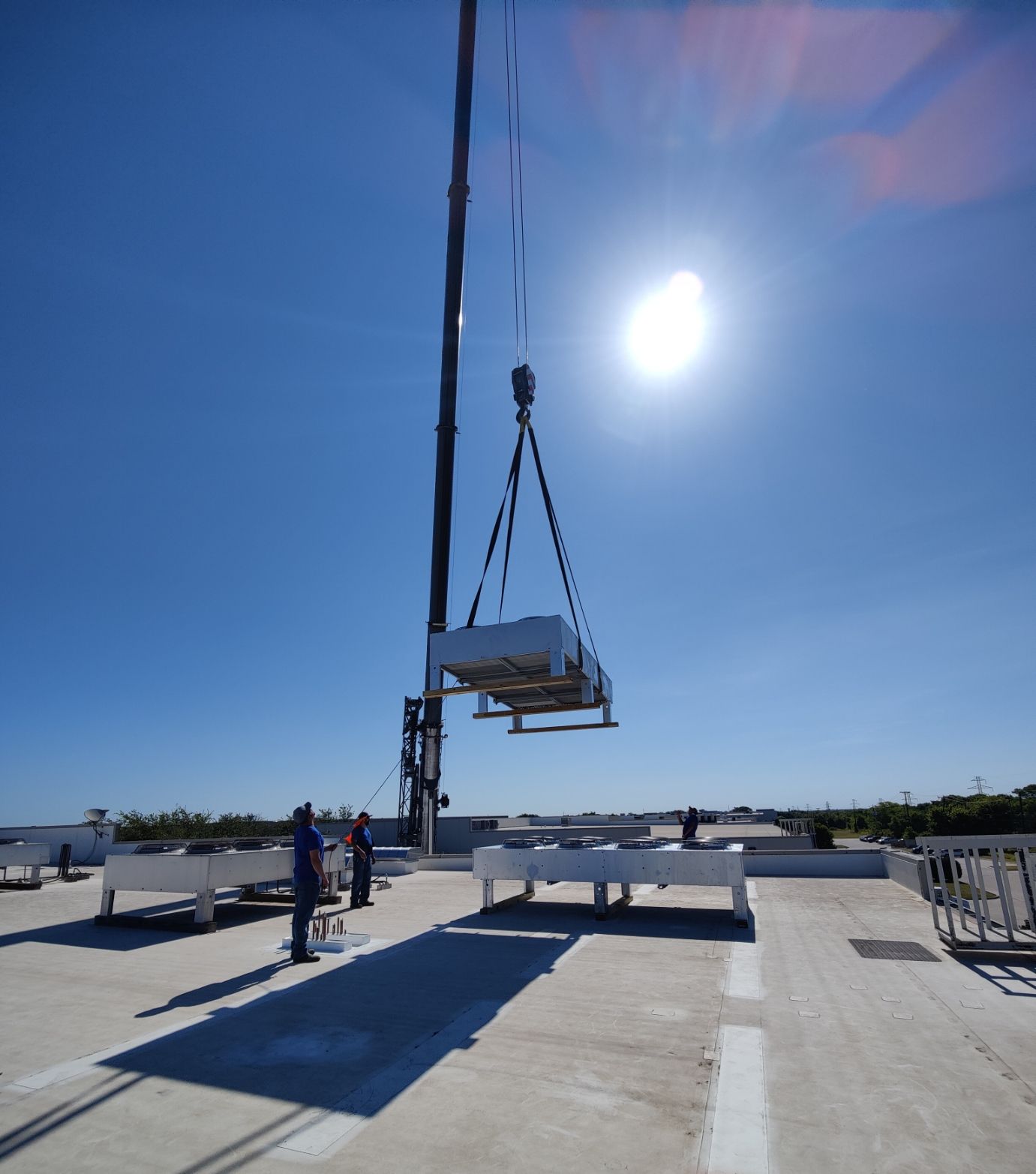 Crane lifting HVAC unit onto a flat rooftop under a bright, sunny sky.