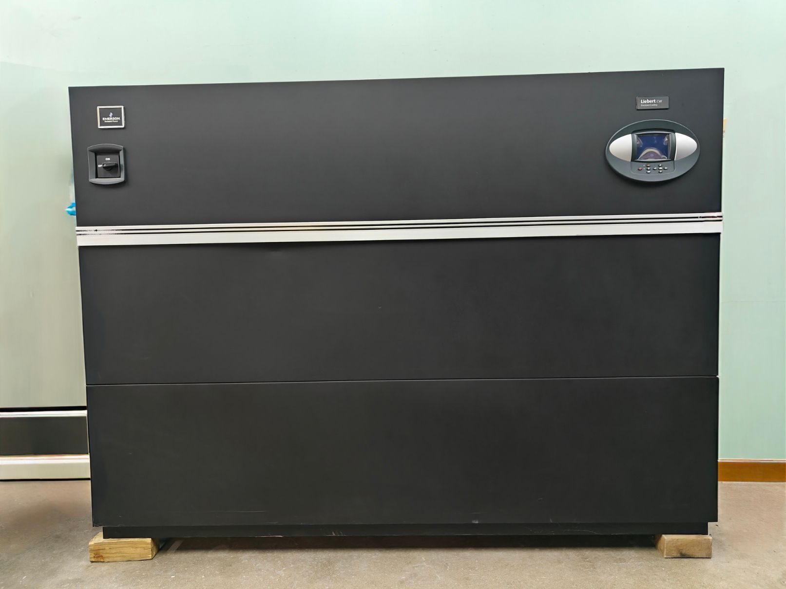 Dark gray industrial-looking machine with control panel, mounted on a wooden pallet, against a pale green wall.