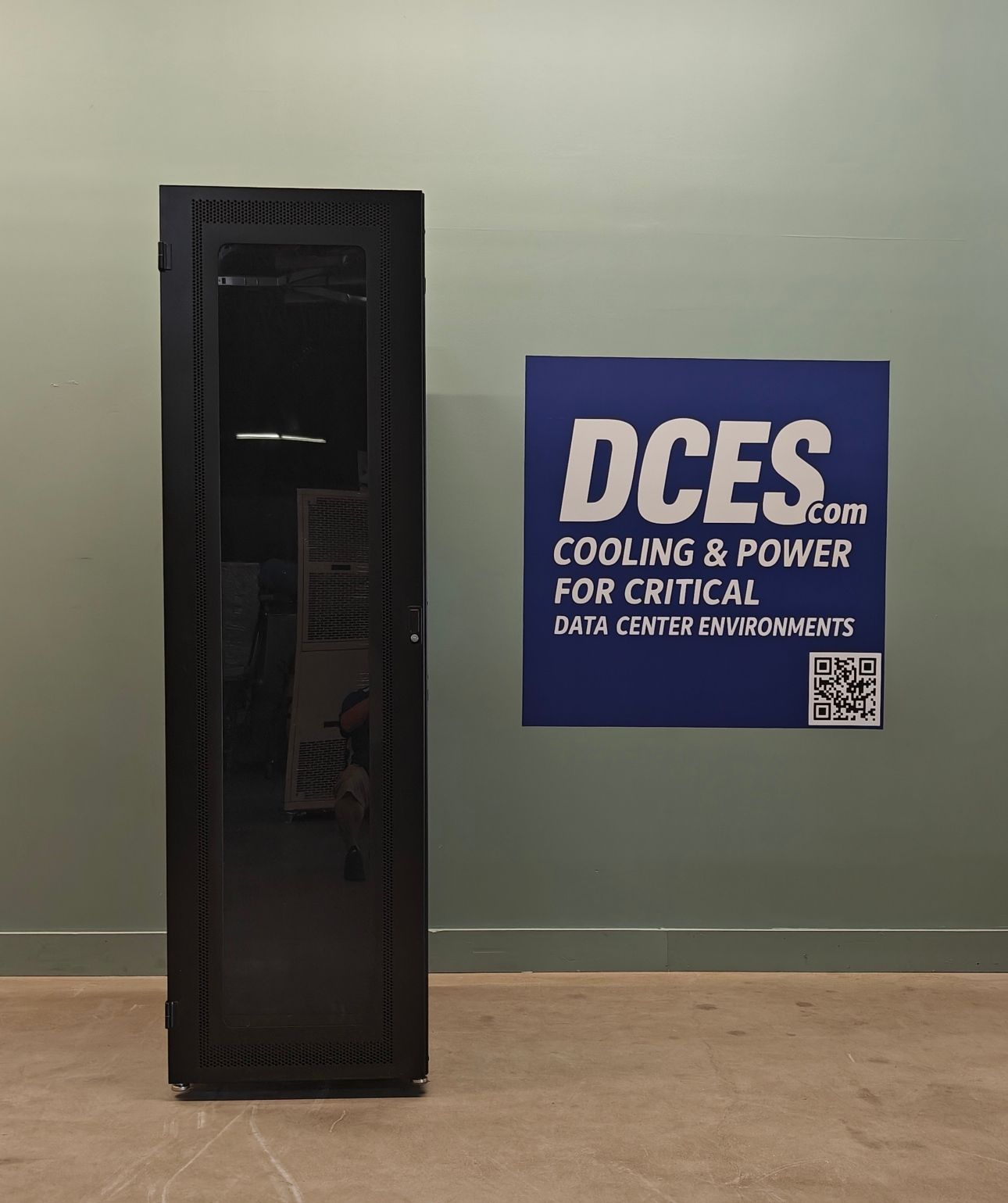 Black server rack next to a blue sign for DCES, in a room with a concrete floor.