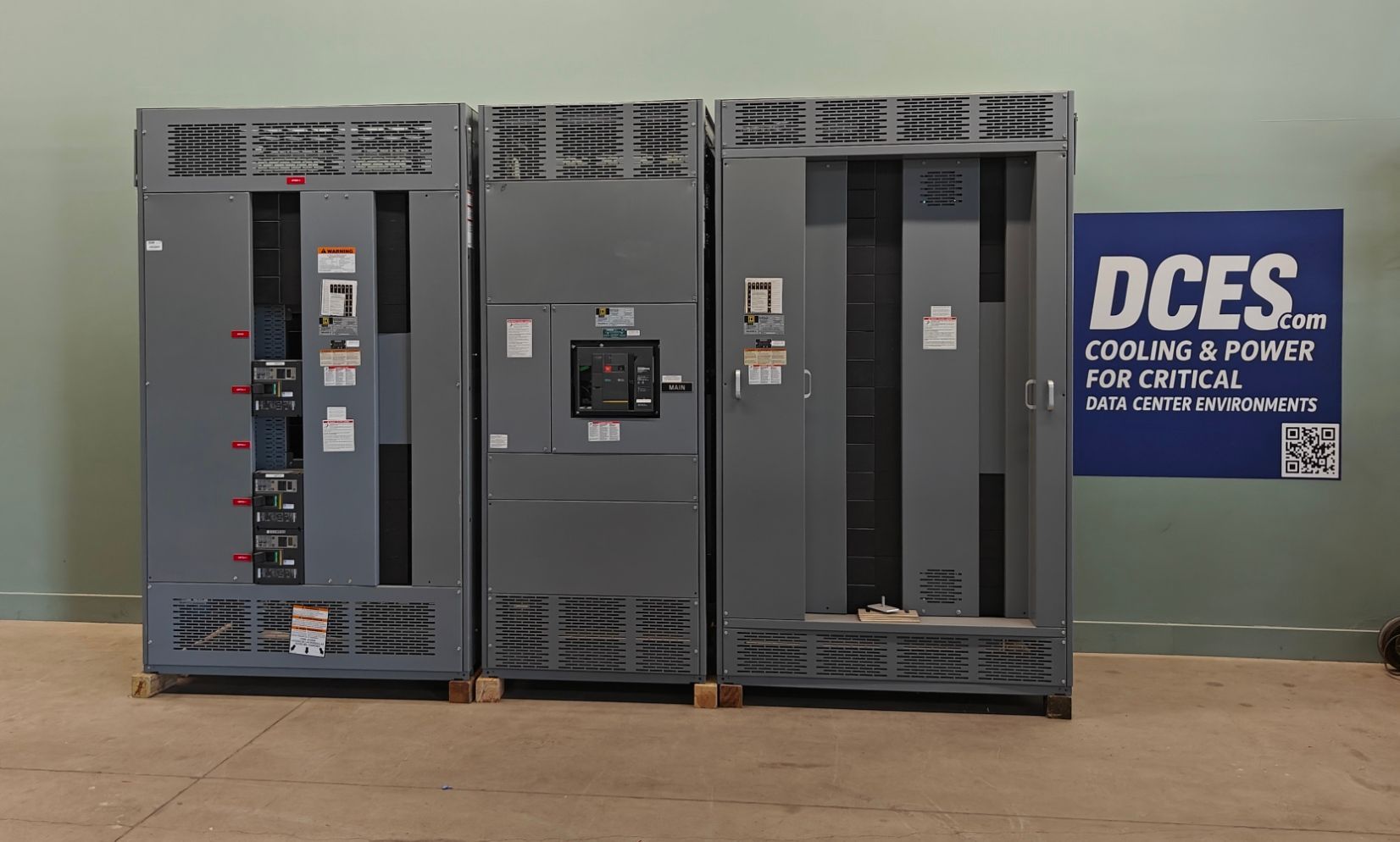 Three gray electrical switchgear cabinets on wooden pallets in a warehouse.