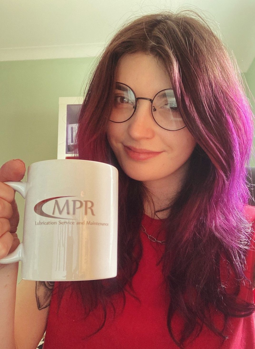 girl with purple hair sitting in office drinking tea out of a white mug with MPR written on it, customised, smiling