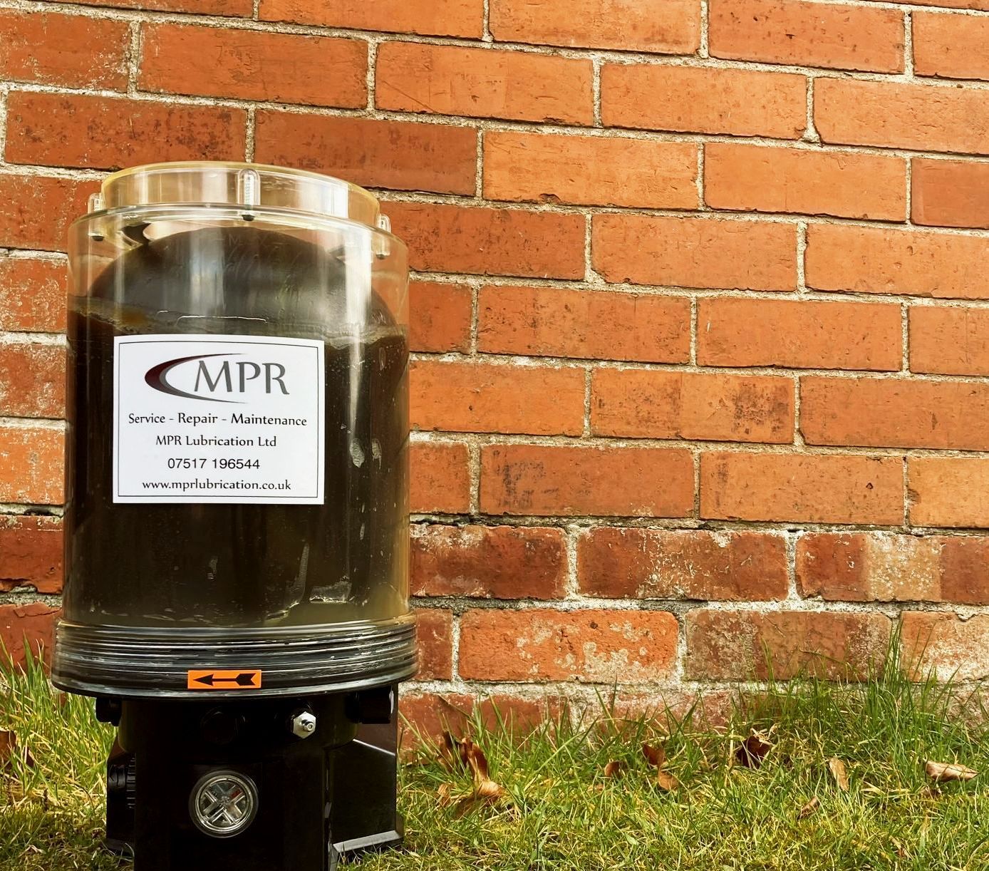 MPR branded automatic lubrication pump sitting on grass in front of brick wall, autolube pump