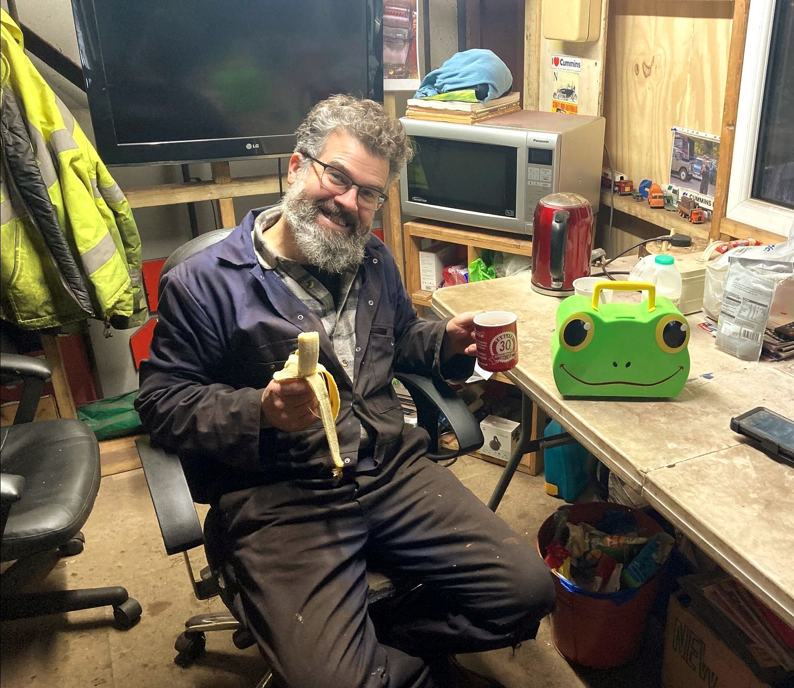 man with beard and glasses in workshop, eating a banana and drinking a cup of tea with his frog shaped lunchbox.