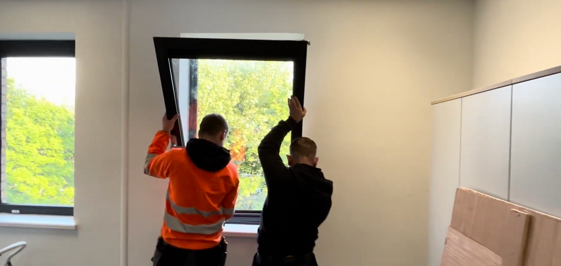 Two workers in orange and black lift a large window frame in a bright room.