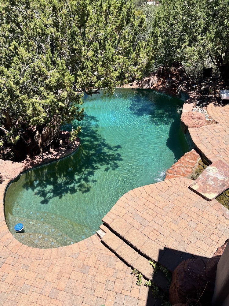 A kidney-shaped swimming pool with teal water surrounded by a reddish stone patio and lush green trees.
