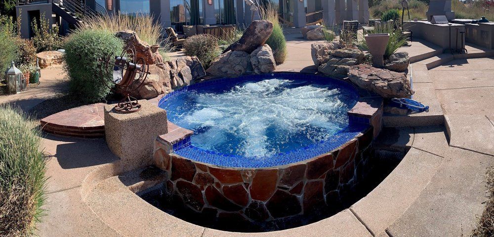 A stone-clad circular hot tub with swirling water sits on a backyard patio, surrounded by desert landscaping and rocks.