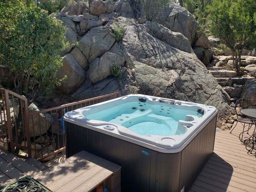 A kidney-shaped swimming pool with teal water surrounded by a reddish stone patio and lush green trees.