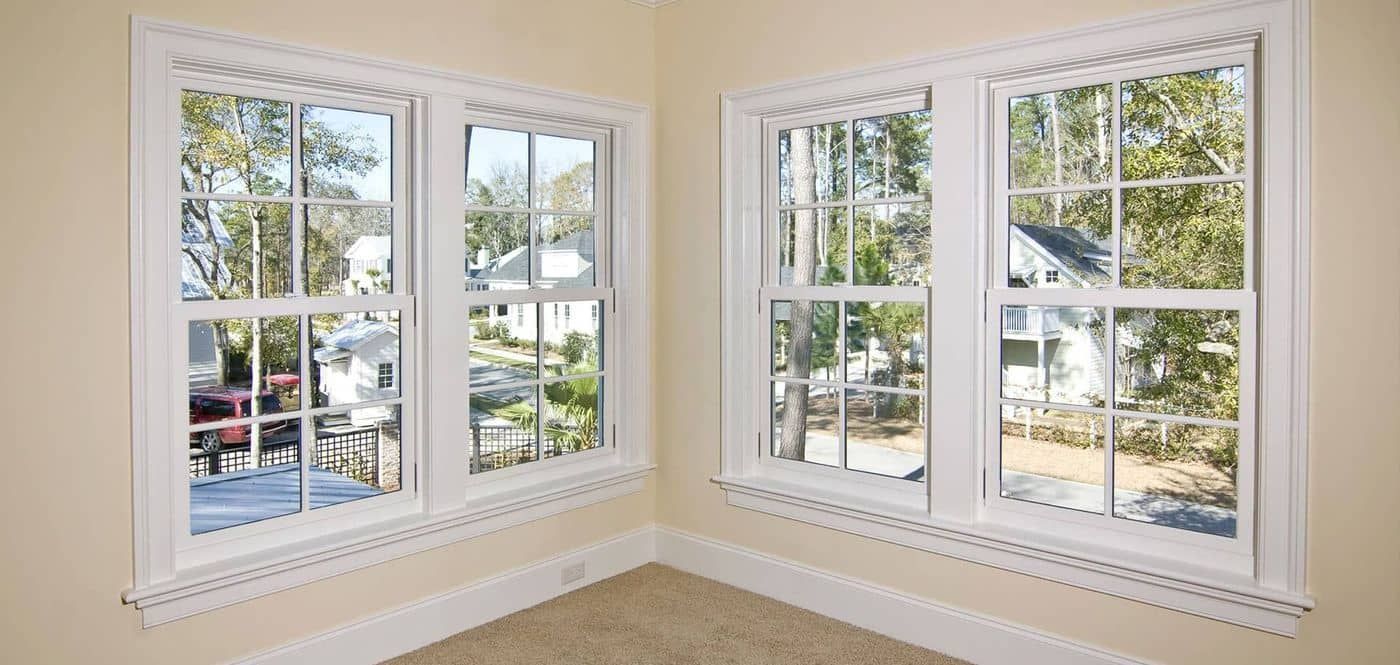 Corner of a room with two white, multi-paned windows overlooking a street with houses and trees.