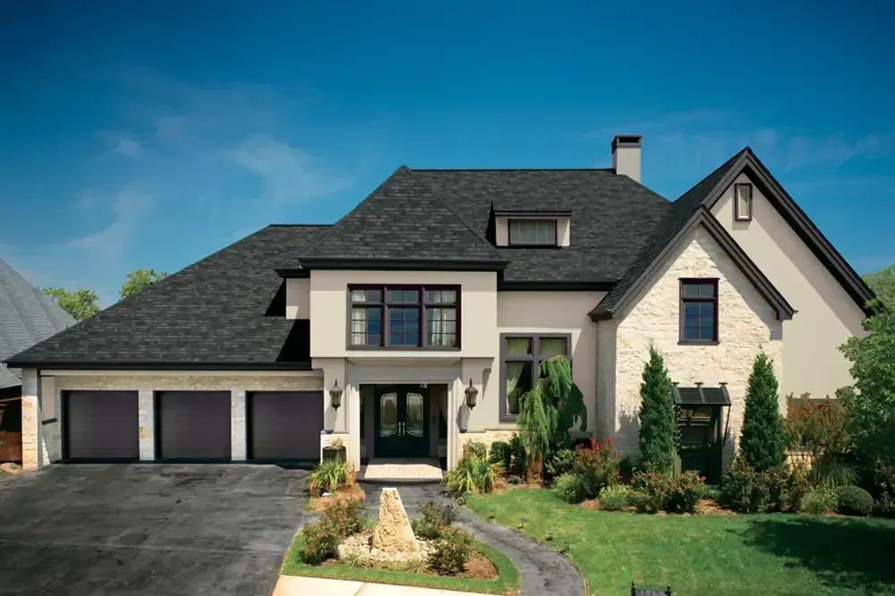 Two-story house with black roof, beige stucco, three-car garage, and green lawn under a blue sky.