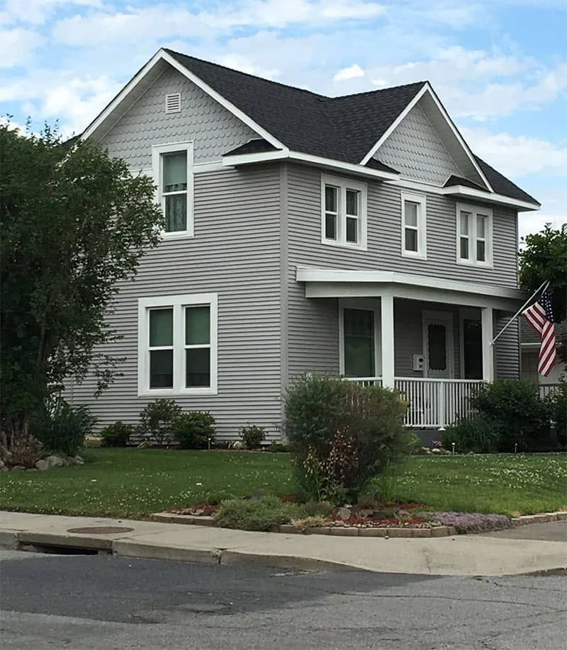 Two-story gray house with white trim, a porch, and an American flag on a grassy corner lot.