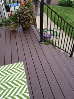 Brown deck with stairs, black railing, and potted plants; green and white patterned rug in foreground.