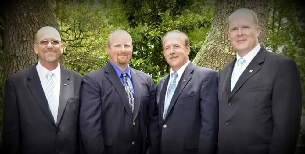 Four men in suits and ties are standing next to each other in front of trees.