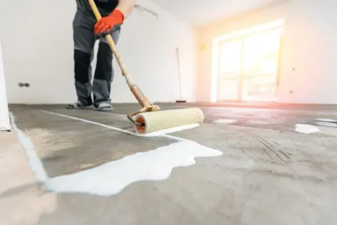 Person applying white paint to a concrete floor with a roller in a bright room