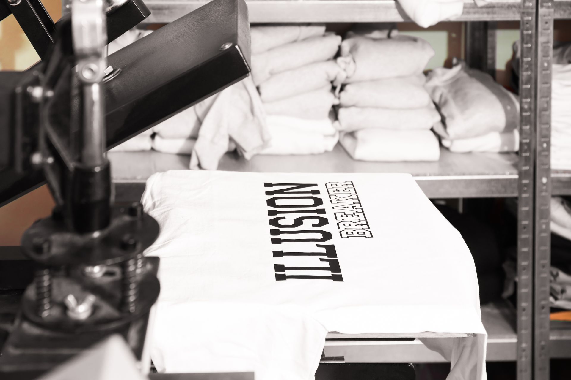 A white T-shirt on a screen-printing press with bold black lettering being printed in a workshop.
