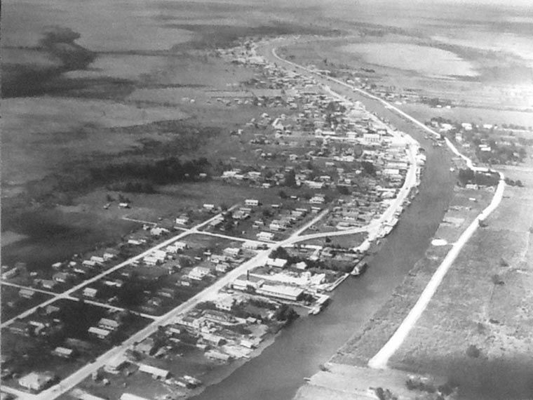 The First Paved Highway. The town's original shell road on the west bank of Bayou Lafourche (on the left in this photo) was paved in concrete in 1936. This road was the predecessor of LA 1, along which the town rapidly developed. On the east bank, which is relatively unsettled, the road remains shelled and abruptly stops at a clearing the right foreground. In the left foreground, with cross-streets paralleling the road, is a subdivision of largely fishermen's homes called Rue Poche Vide, or 