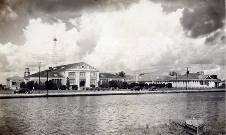 Iconic Structures of Early Town. (c) The Golden Meadow High School building in the late 1930s, with a drilling derrick in the background and an overturned chaland across the bayou in the foreground. Photo courtesy of the Golden Meadow Library Historical Center.