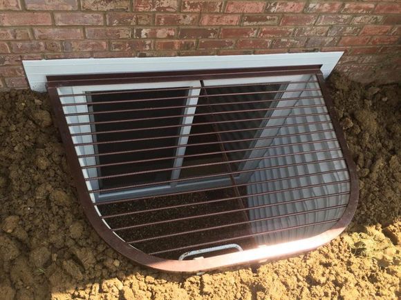 Basement window well with metal grate below a brick wall, partly filled with dirt