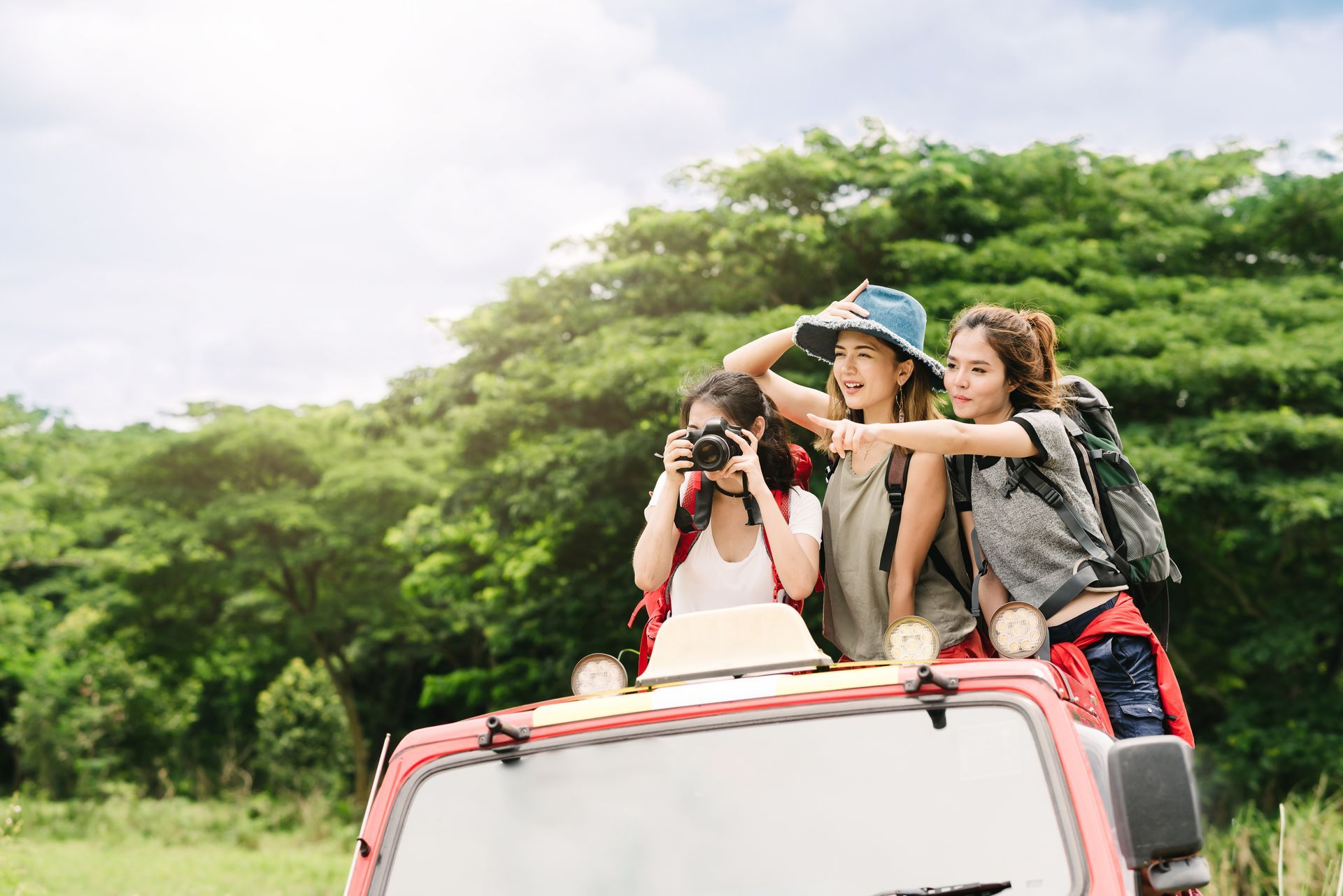 Three women on a red vehicle, looking at scenery. One takes photos, another points, and the third wears a hat. Lush green trees in background.