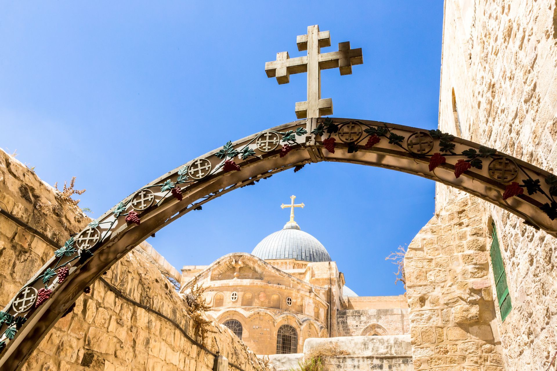Archway with a stone cross, leading to a church with a blue dome in Jerusalem.