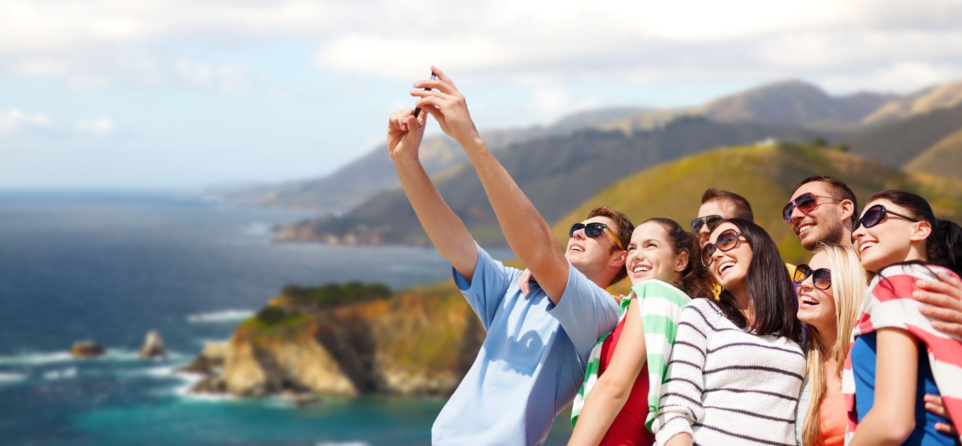 Group of people taking a selfie outdoors with a scenic ocean and mountain backdrop.