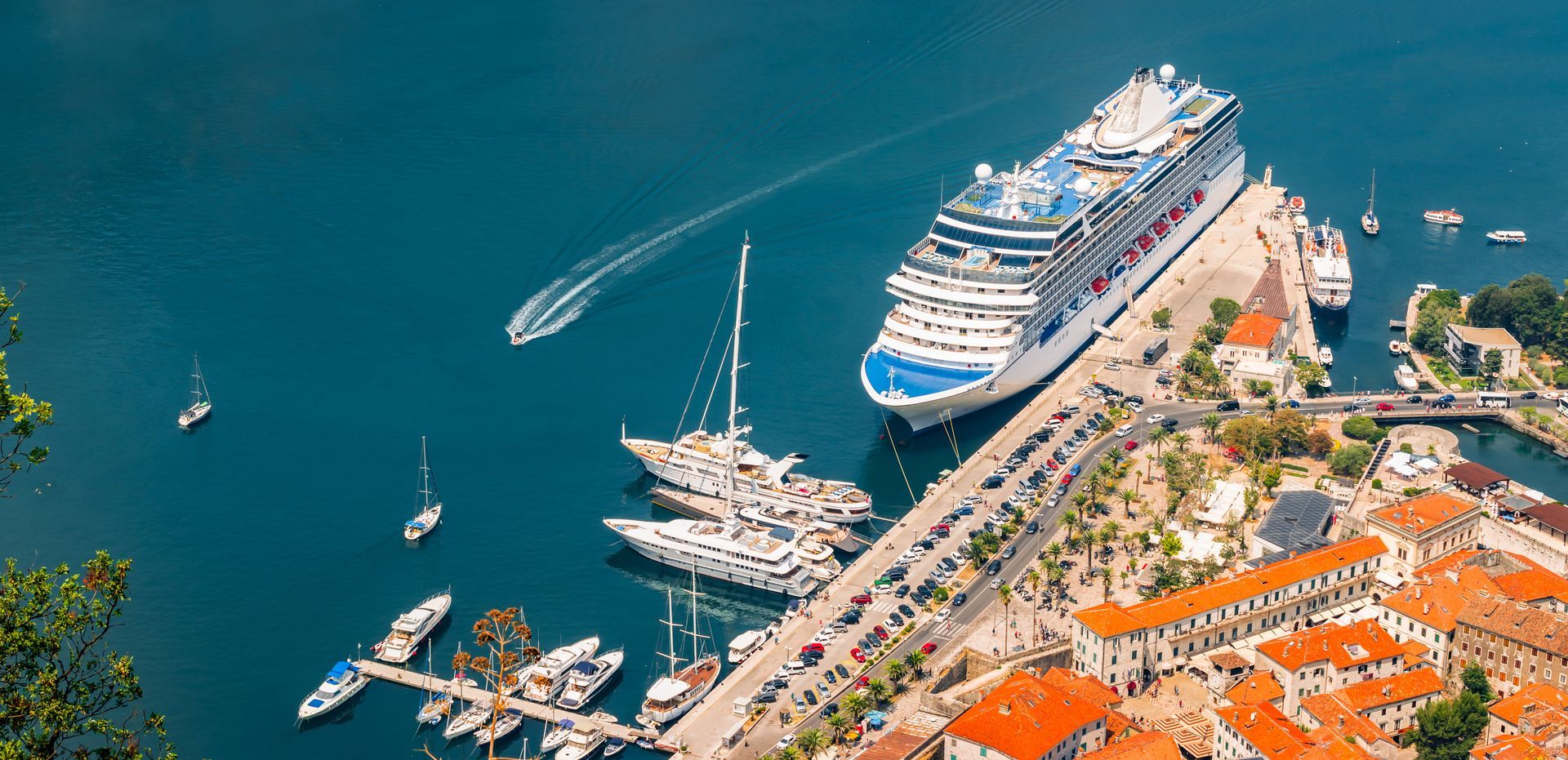 Aerial view of a cruise ship docked in a harbor, with sailboats and town with orange rooftops.