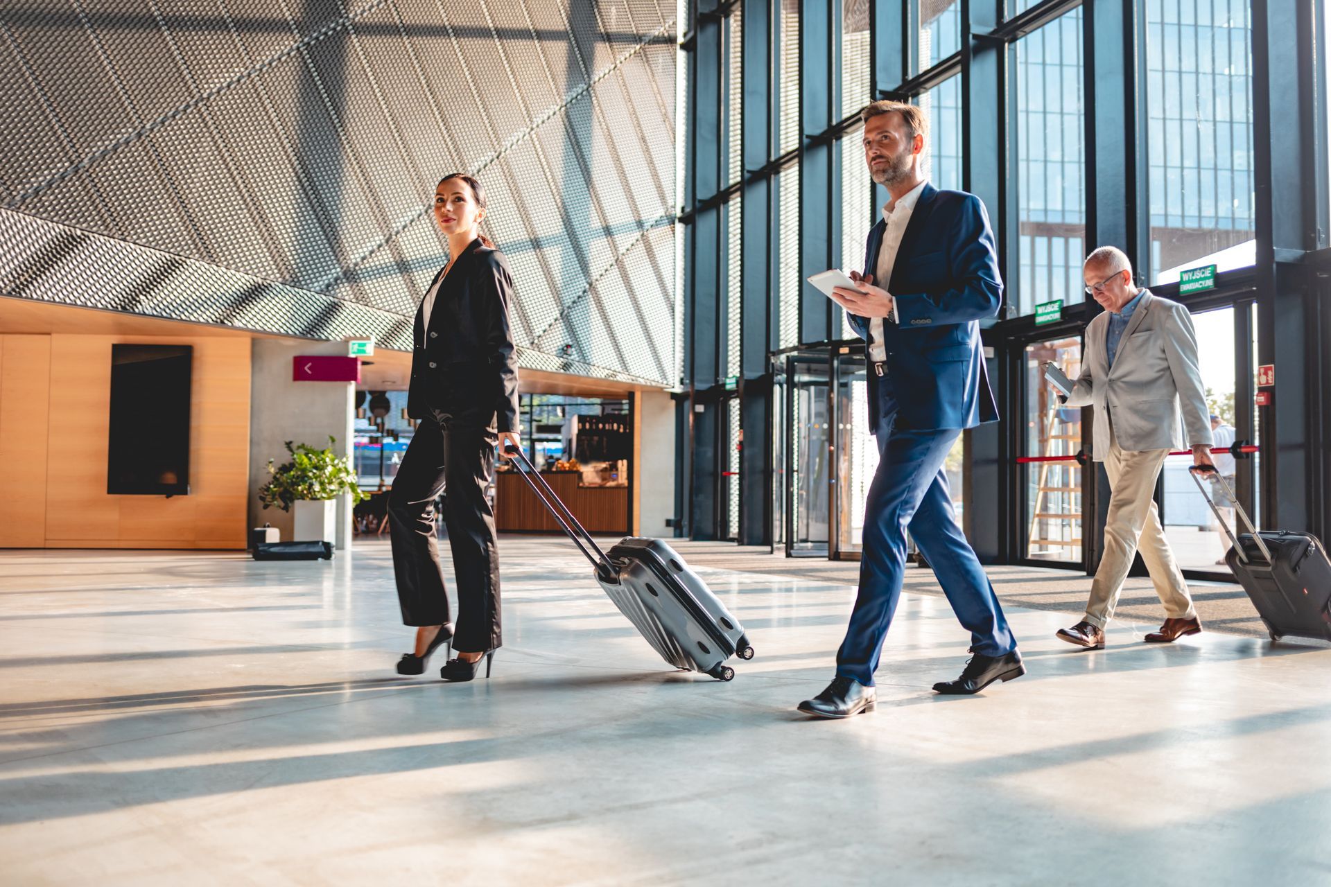 Three people with rolling luggage walk through a modern building lobby with glass doors.