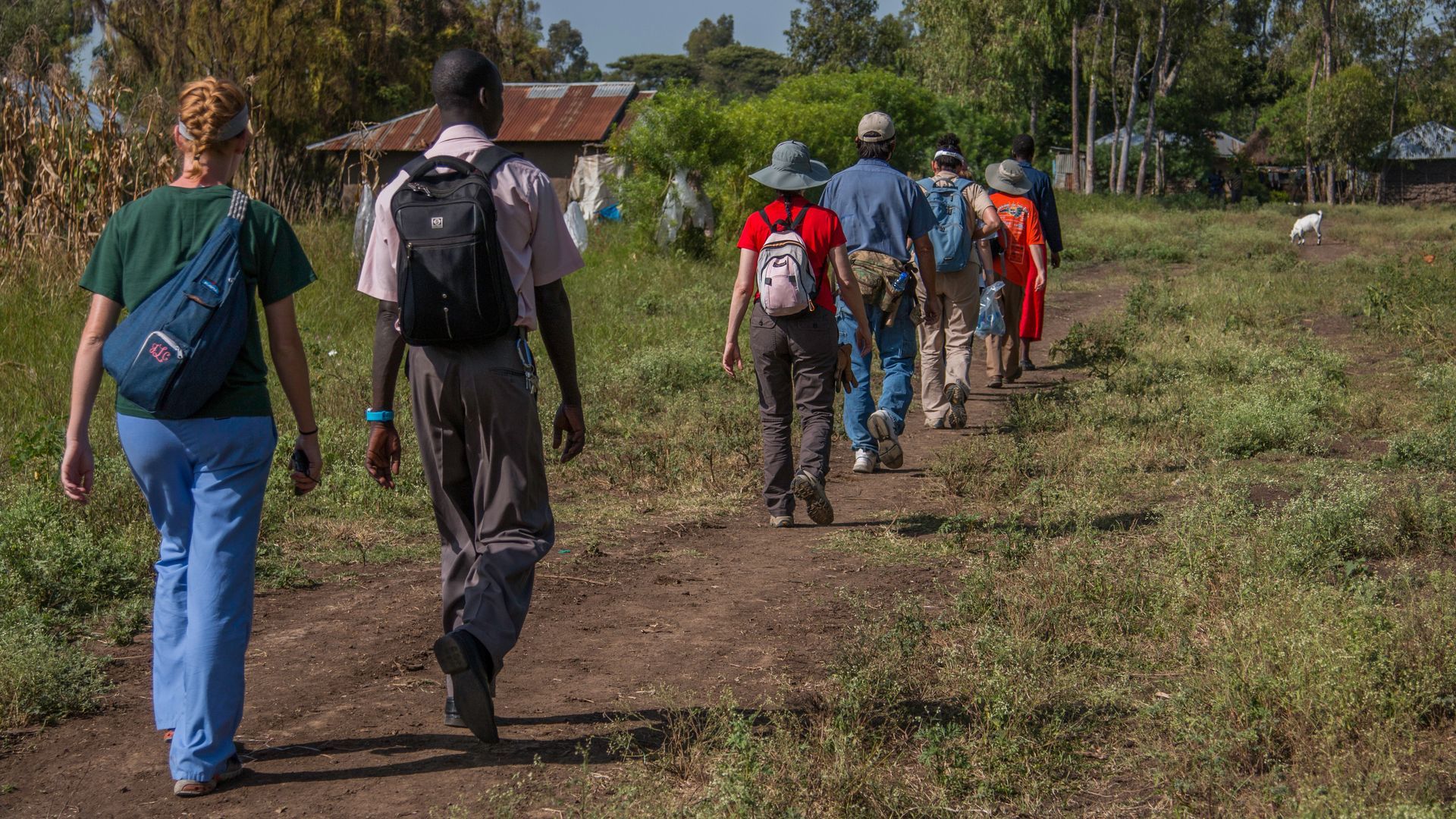 Group of people walking on a dirt path in a rural area, carrying backpacks.