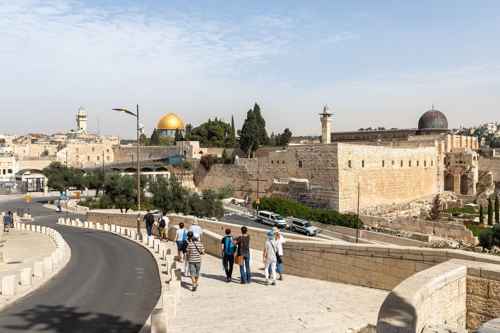 Jerusalem cityscape with the Dome of the Rock and a stone wall, people walking on a walkway, bright daylight.