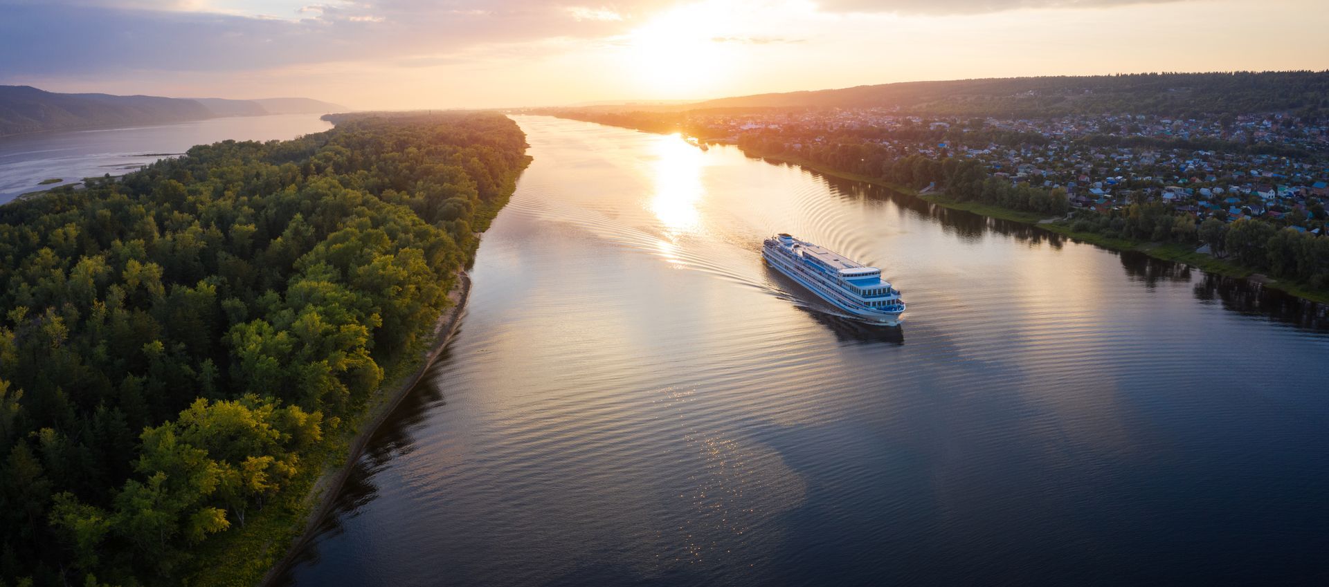 River cruise ship sailing on a wide river at sunset. A lush green island is on the left.