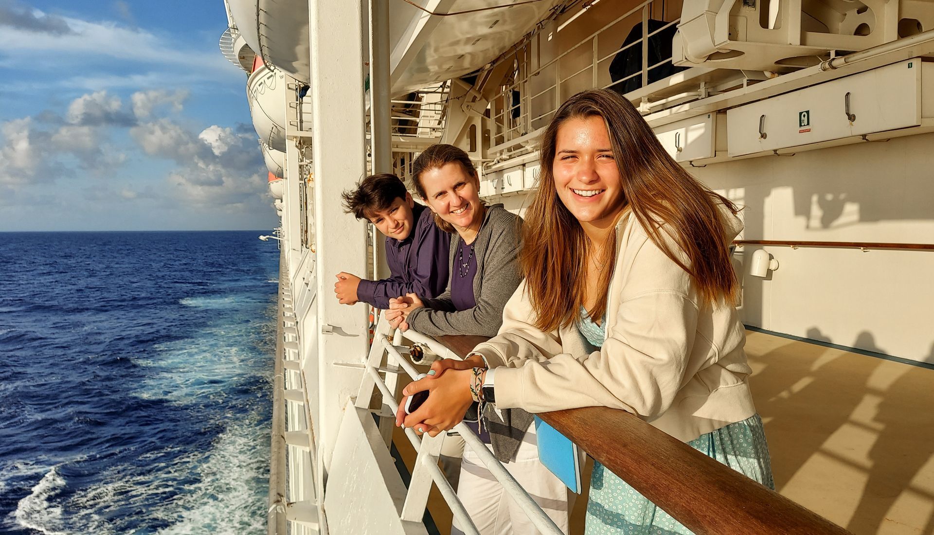 Three people smiling on a cruise ship deck, looking at the ocean.