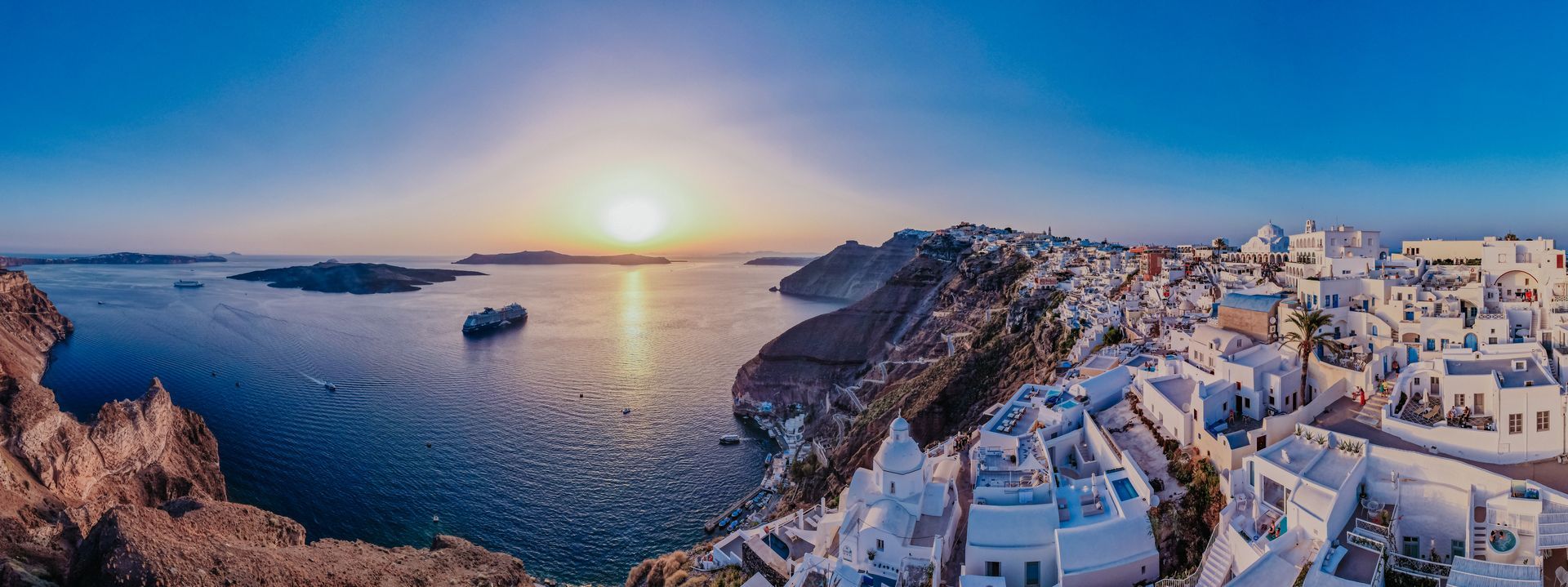 Panoramic view of Santorini, Greece at sunset: white buildings on a cliff, blue sea, and a setting sun.